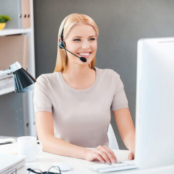 Frau mit Headset, die lächelnd am Schreibtisch sitzt und am Computer arbeitet.